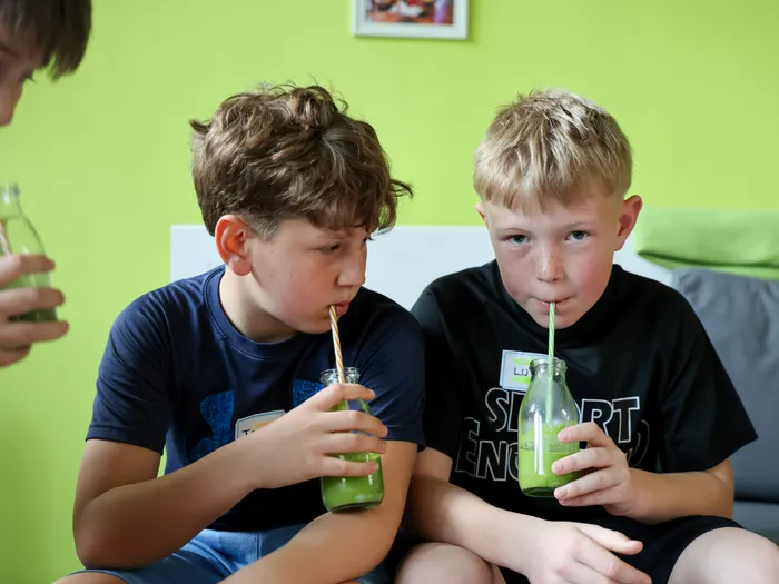 Zwei Jungen trinken aus Flaschen mit einem Strohhalm einen grünen Smoothie. Im Hintergrund ist ein weiterer Junge zu sehen, der ebenfalls ein Getränk hält.