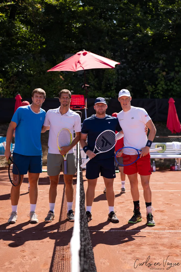 Vier Tennisspieler stehen hinter einem Netz auf einem Sandplatz und halten ihre Schläger.
