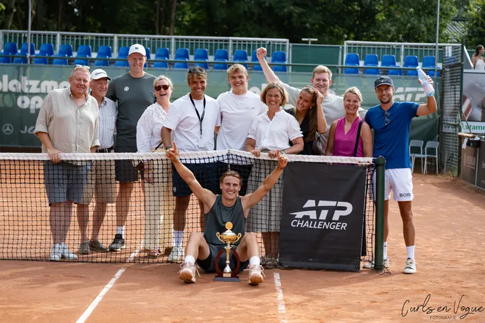 Tom Gentzsch sitzt mit erhobenen Armen vor seinem Pokal auf einem Sandplatz, während eine Gruppe von Menschen hinter dem Tennisnetz feiert.