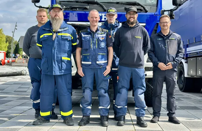 Sechs Helfer des Technischen Hilfswerks in blauer Arbeitskleidung stehen vor einem blauen Einsatzfahrzeug.