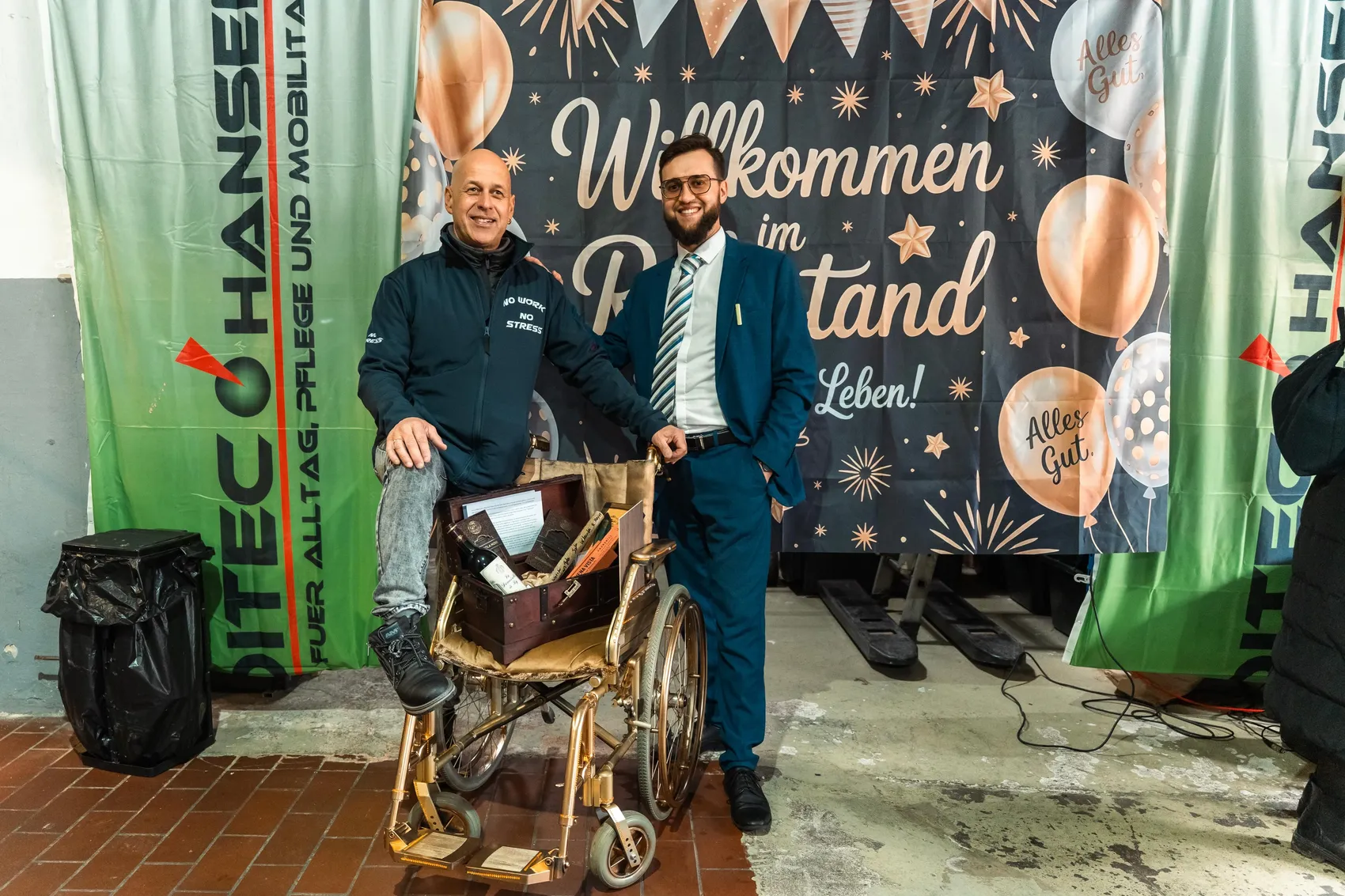 Zwei lächelnde Männer stehen vor einem Rollstuhl und einem Banner mit der Aufschrift „Willkommen im Tandem Leben!“. Im Hintergrund ist ein Werbebanner für das Sanitätshaus Meditec Nachrodt zu sehen.