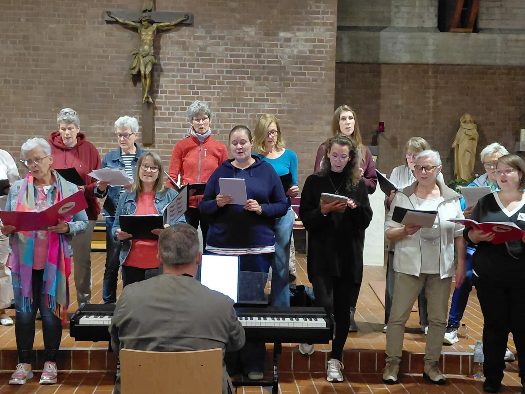 Chor singt in einer Kirche, begleitet von einem Keyboardspieler. Mehrere Personen halten Notenblätter in den Händen.