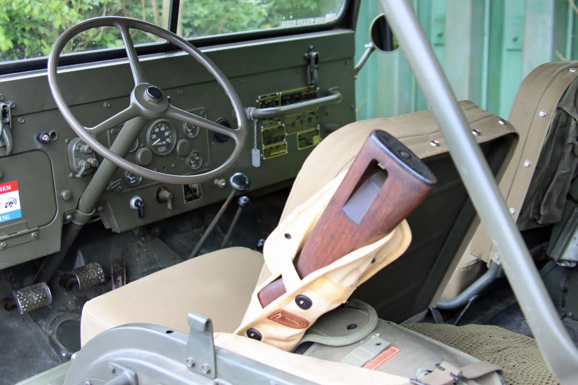 Innenansicht eines olivgrünen Oldtimer-Jeeps mit Lenkrad und beigem Sitz, auf dem ein Gewehr liegt.