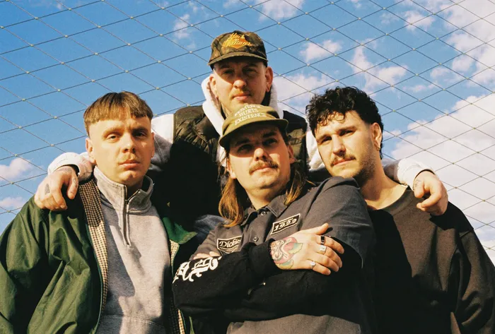 Die vier Mitglieder der Band Poppy Wizard posieren vor einem Maschendrahtzaun unter blauem Himmel.