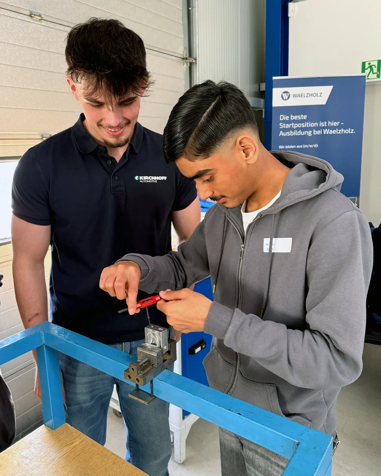 Ein Ausbilder erklärt einem Schüler die Handhabung einer roten Zange an einem Metallteil während einer praktischen Übung in einer Werkstatt.