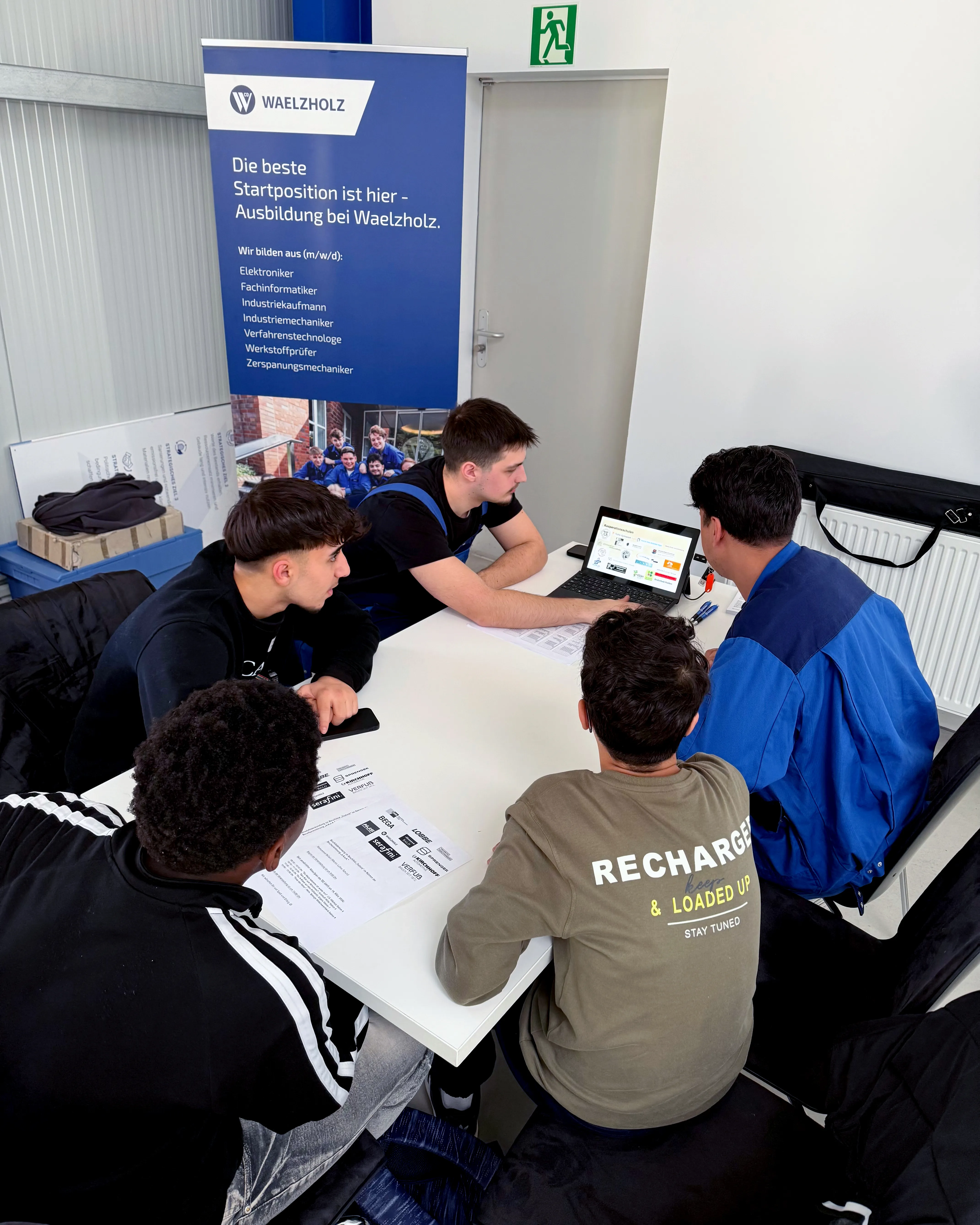 Vier junge Menschen sitzen an einem Tisch und schauen auf einen geöffneten Laptop. Im Hintergrund ein Werbebanner für Ausbildungsberufe bei Waelzholz.