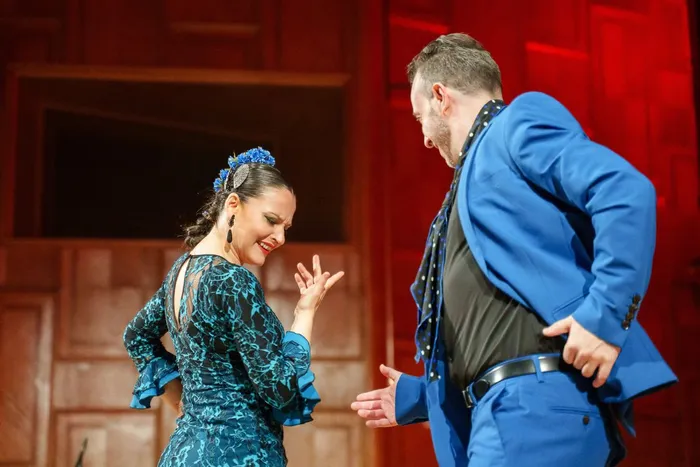Eine Frau in einem blau gemusterten Kleid und ein Mann in einem blauen Anzug tanzen Flamenco auf einer Bühne.
