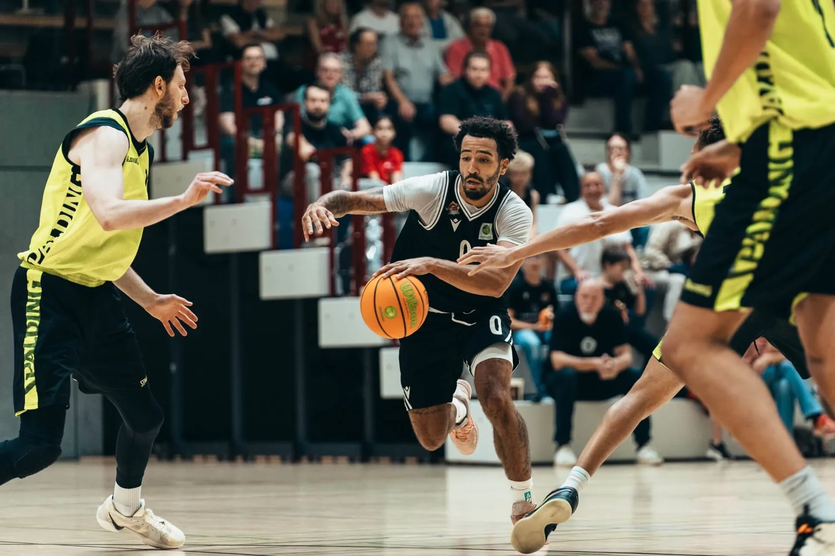 Ein Basketballspieler in einem weiß-schwarzen Trikot dribbelt den Ball, während er von Spielern in gelben Trikots verteidigt wird.