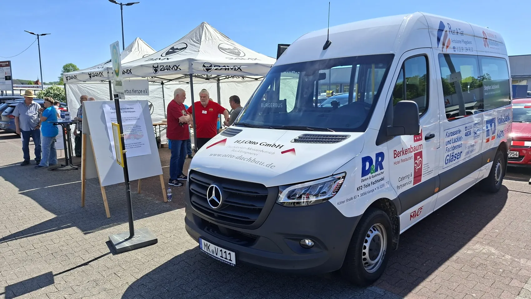 Weißer Transporter mit Aufschrift „DR Berkenbaum Hotel Restaurant“ und Logos von Partnern steht auf einem Parkplatz vor mehreren Pavillons mit Werbematerialien.