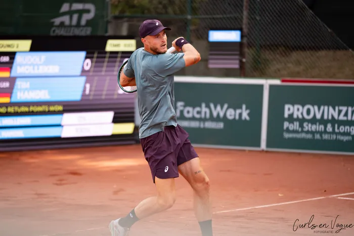 Rudolf Molleker schlägt auf einem Sandplatz einen Tennisball. Er trägt ein graues T-Shirt, dunkle Shorts und eine Kappe.
