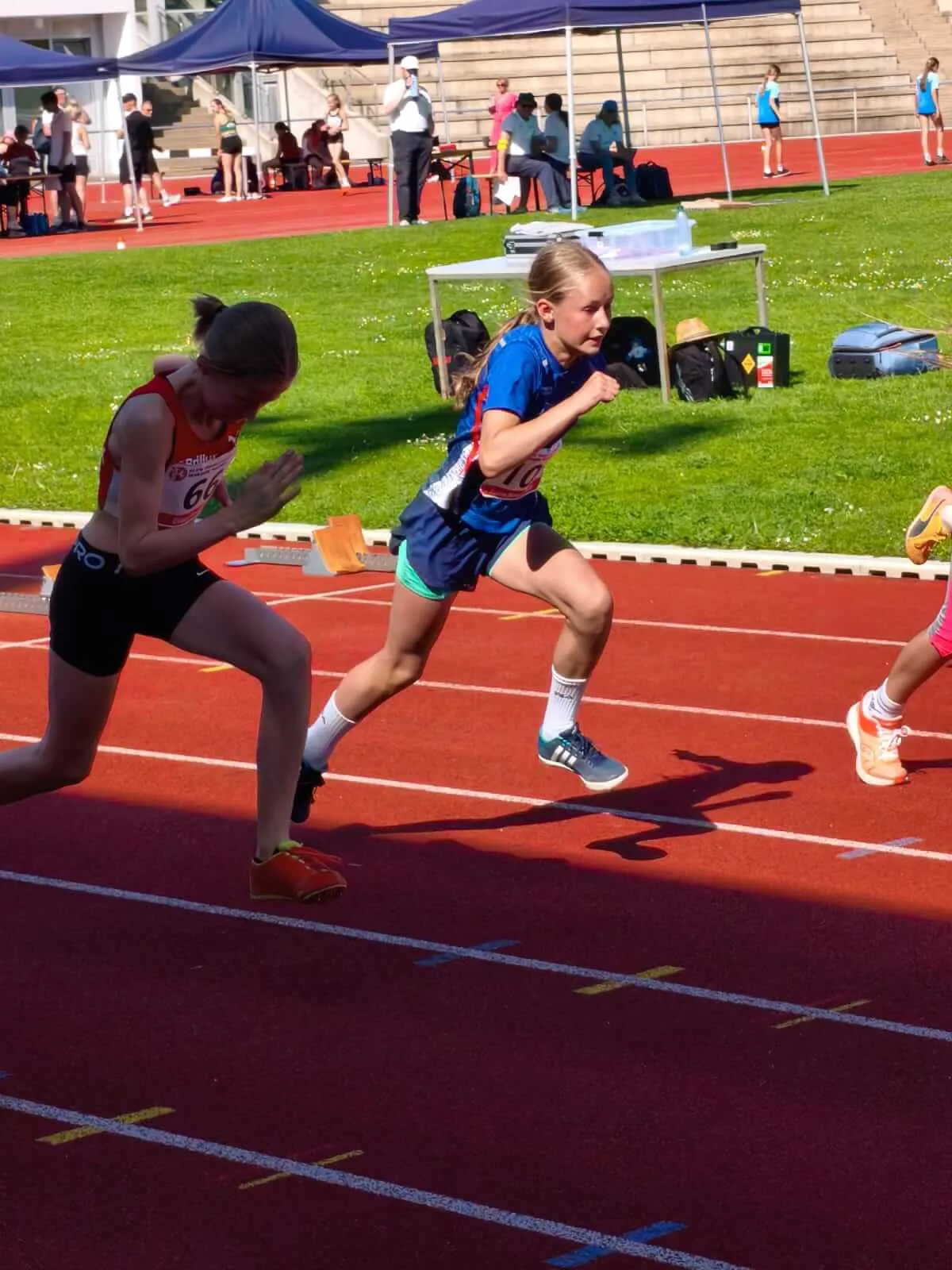 Zwei junge Athletinnen in Laufbekleidung starten auf einer roten Tartanbahn bei einem Wettkampf, im Hintergrund Zuschauer und blaue Zelte.