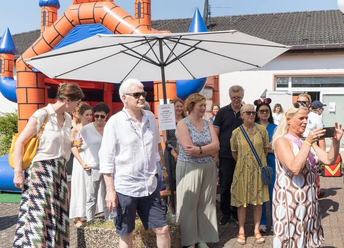 Menschen stehen vor einer aufblasbaren Burg und beobachten ein Ereignis. Im Hintergrund ein Gebäude und ein Sonnenschirm.