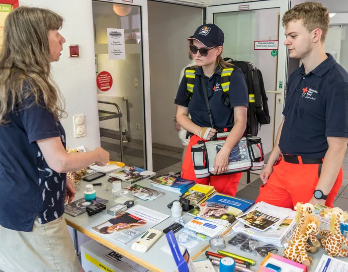 Zwei ehrenamtliche Helfer der Feuerwehr und des Deutschen Roten Kreuzes informieren an einem Infostand eine Besucherin. Auf dem Tisch liegen Informationsmaterialien und Erste-Hilfe-Utensilien.