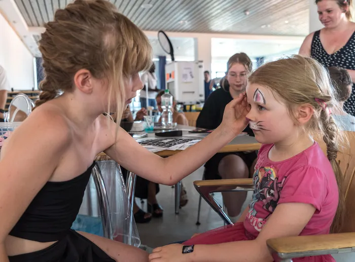 Eine Frau bemalt einem jungen Mädchen ein Kätzchen auf die Wange. Im Hintergrund sind weitere Personen und eine helle, offene Raumgestaltung zu sehen.