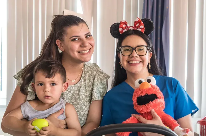 Zwei Frauen und zwei Kinder posieren lächelnd für ein Foto. Ein Kind sitzt in einem Autositz, während ein anderes von einer Frau gehalten wird. Eine der Frauen trägt ein Stirnband mit Micky-Maus-Ohren und hält eine Elmo-Puppe.