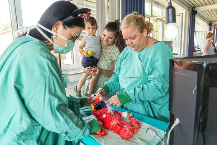 Ärztinnen und Ärzte bereiten ein rotes Stofftier auf einer Liege vor, während ein kleines Kind und seine Mutter zusehen. Im Hintergrund sind weitere Personen und ein heller Raum erkennbar.