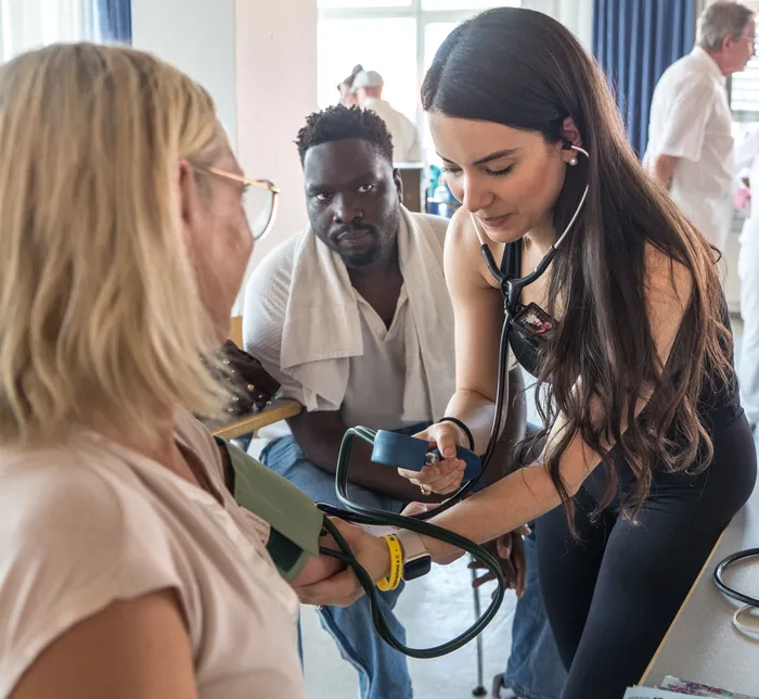 Eine junge Ärztin misst einer älteren Patientin den Blutdruck, während ein Mann im Hintergrund aufmerksam zusieht. Die Szene spielt in einem hellen Raum, möglicherweise in einer Arztpraxis oder Klinik.