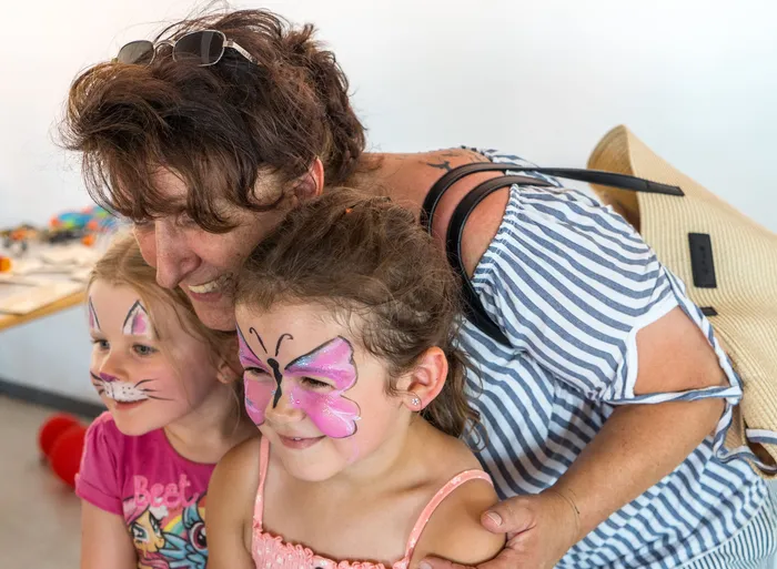 Eine Frau mit lockigem, braunem Haar umarmt zwei Mädchen. Beide Kinder haben Gesichtsbemalungen: eines eine Katze, das andere einen Schmetterling. Im Hintergrund ist ein Tisch mit Spielzeug und Farbe zu sehen.