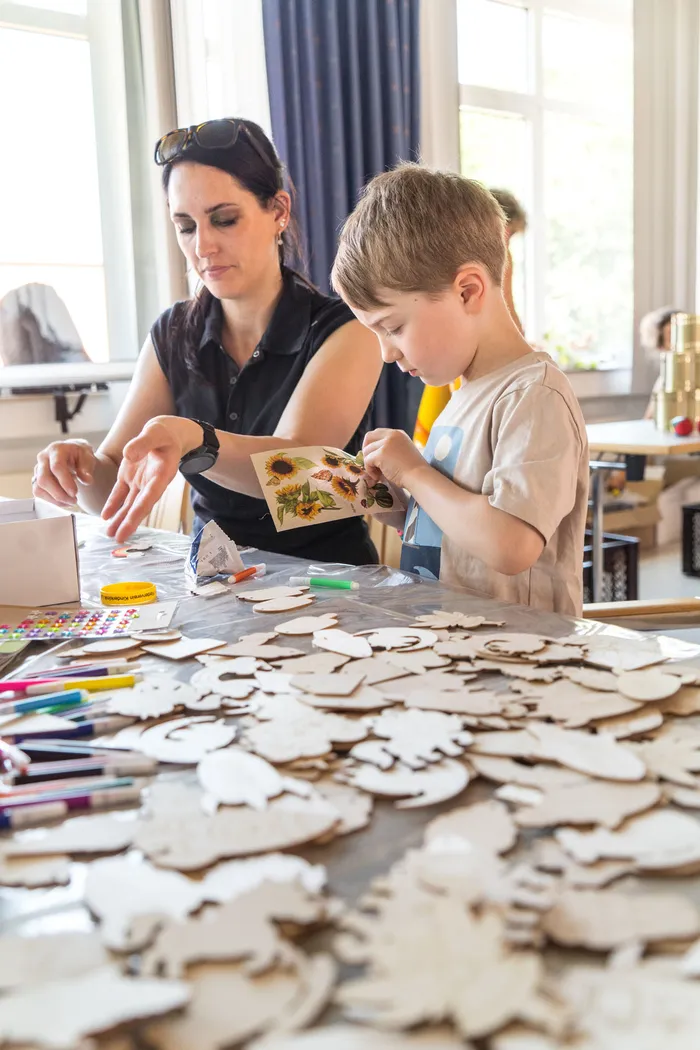 A woman and a boy are working on a craft project at a table covered with wooden shapes. The boy is holding a card with sunflowers, while the woman assists him.