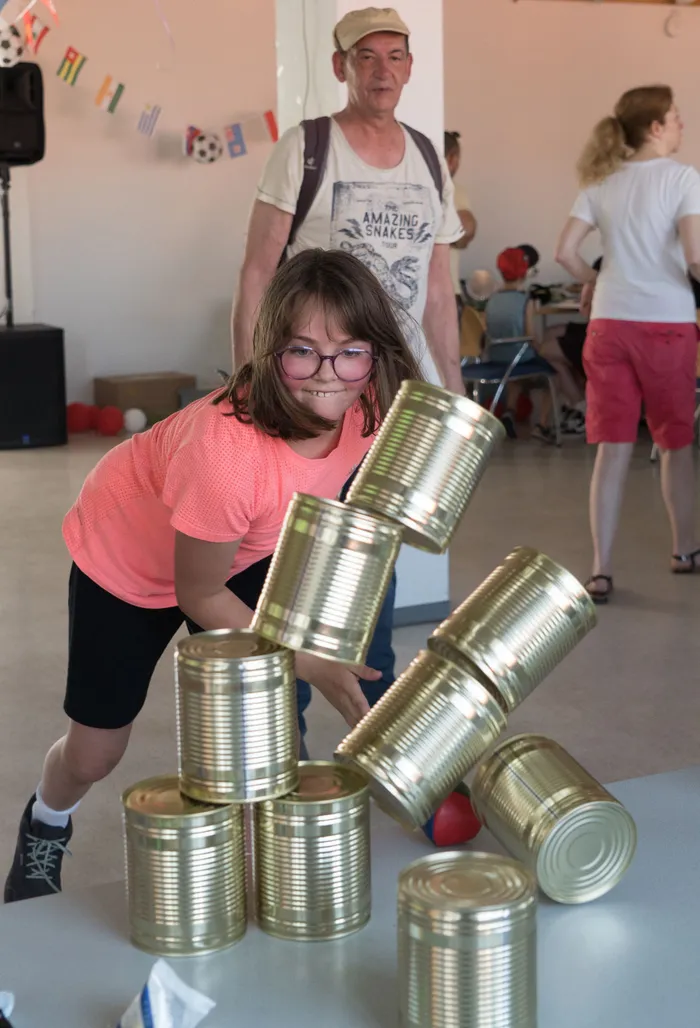 Ein Mädchen mit Brille baut konzentriert eine Pyramide aus Konservendosen auf. Im Hintergrund steht ein Mann und eine weitere Person ist teilweise zu sehen.