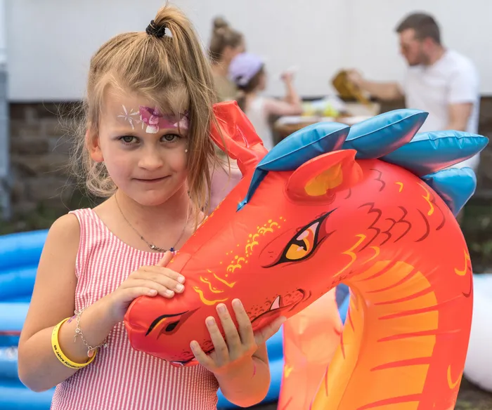 Ein Mädchen mit Gesichtsbemalung hält einen aufblasbaren Drachen vor sich. Im Hintergrund sind weitere Kinder und ein Pool zu sehen.