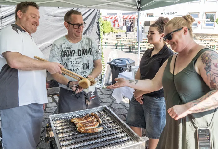 Ein Mann grillt Würstchen vor einem Stand mit dem Aufdruck „Camp David“. Zwei Frauen nehmen am Stand Essen entgegen. Im Hintergrund sind Tische und weitere Personen zu sehen.