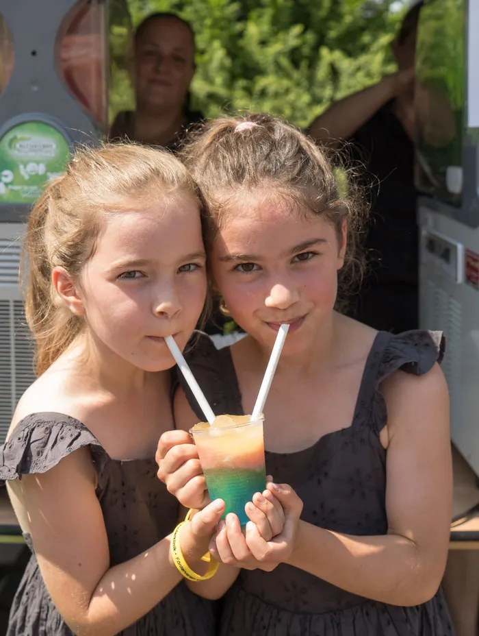Zwei Mädchen teilen sich einen farbigen Drink mit Strohhalmen. Im Hintergrund ist ein Fahrzeug zu sehen, vermutlich ein Foodtruck oder ähnliches.
