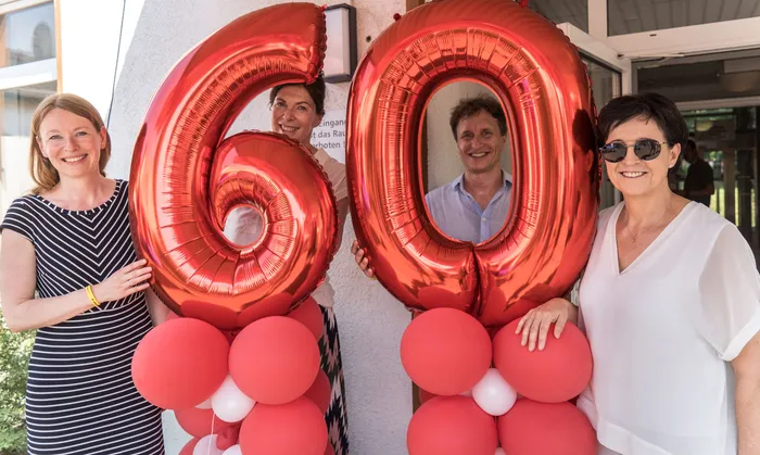 Vier Personen stehen vor einer hellen Fassade und halten rote Folienballons in Form der Ziffern 6 und 0. Sie lächeln in die Kamera und feiern den 60. Geburtstag der Kinderklinik Lüdenscheid.