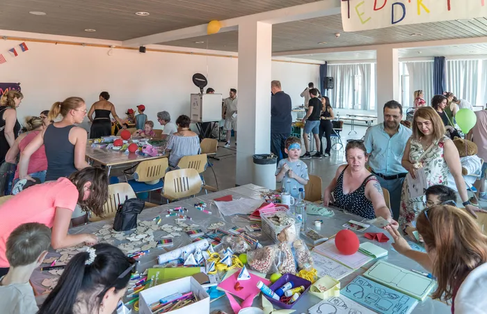 Menschen sitzen und stehen in einem hellen Raum um Tische, an denen gebastelt wird. Im Hintergrund sind weitere Personen und eine Bühne zu sehen.