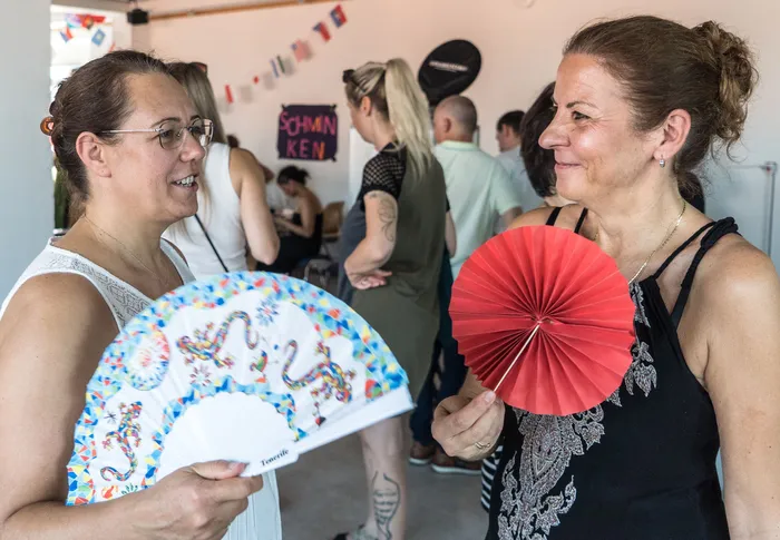 Zwei lächelnde Frauen halten Fächer in der Hand, eine mit einem bunten Drachenmuster, die andere rot. Im Hintergrund sind weitere Menschen auf einer Geburtstagsfeier zu sehen, mit Girlanden und der Aufschrift „Schminken“.
