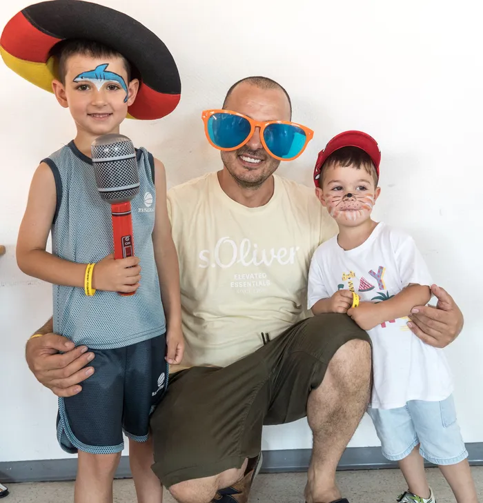 Ein Mann sitzt mit zwei Kindern. Der Mann trägt eine hellbeige T-Shirt und eine große orangefarbene Sonnenbrille, während die Kinder mit Gesichtsbemalung und Hüten geschmückt sind.