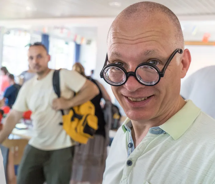Ein lächelnder Mann mit Glatze und großer Brille blickt in die Kamera. Im Hintergrund sind weitere Personen in einem hellen Raum zu sehen.