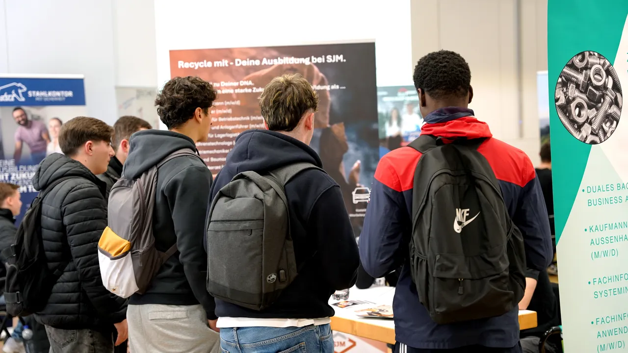 Eine Gruppe junger Menschen steht auf einer Messe oder einem Karrieretag vor einem Infostand und liest Informationen über Ausbildungsangebote bei SJM.