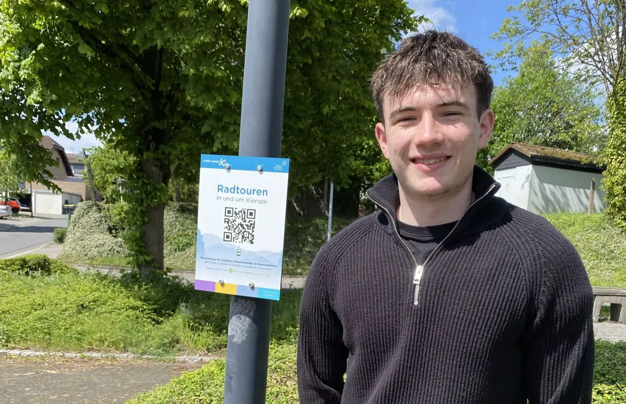 Jüngerer Mann mit Jacke lächelt vor einem Schild mit QR-Code für Radtouren in und um Kierspe.