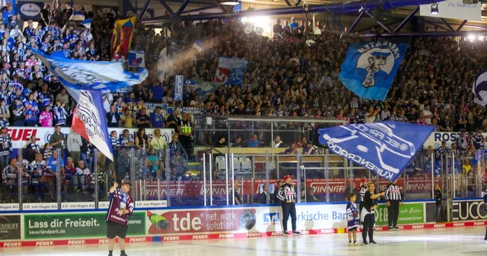 Zuschauer auf den Rängen einer Eishockeyhalle jubeln und schwenken zahlreiche Fanfahnen. Im Vordergrund stehen Spieler auf dem Eis, bereit für den Spielbeginn.
