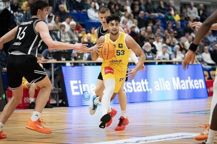 Ein Basketballspieler im gelben Trikot dribbelt den Ball, während ein Gegenspieler in Schwarz-Weiß ihn bedrängt.