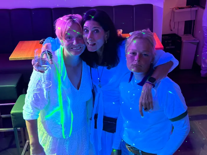 Drei Frauen posieren lächelnd bei der „Party à la Wimbledon“ im Tennisclub Halver unter farbigem Partylicht.