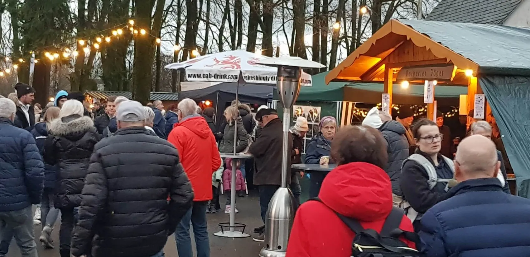 Menschenmenge vor einem kleinen Holzstand mit Lichterketten unter Bäumen bei einer Veranstaltung im Freien.