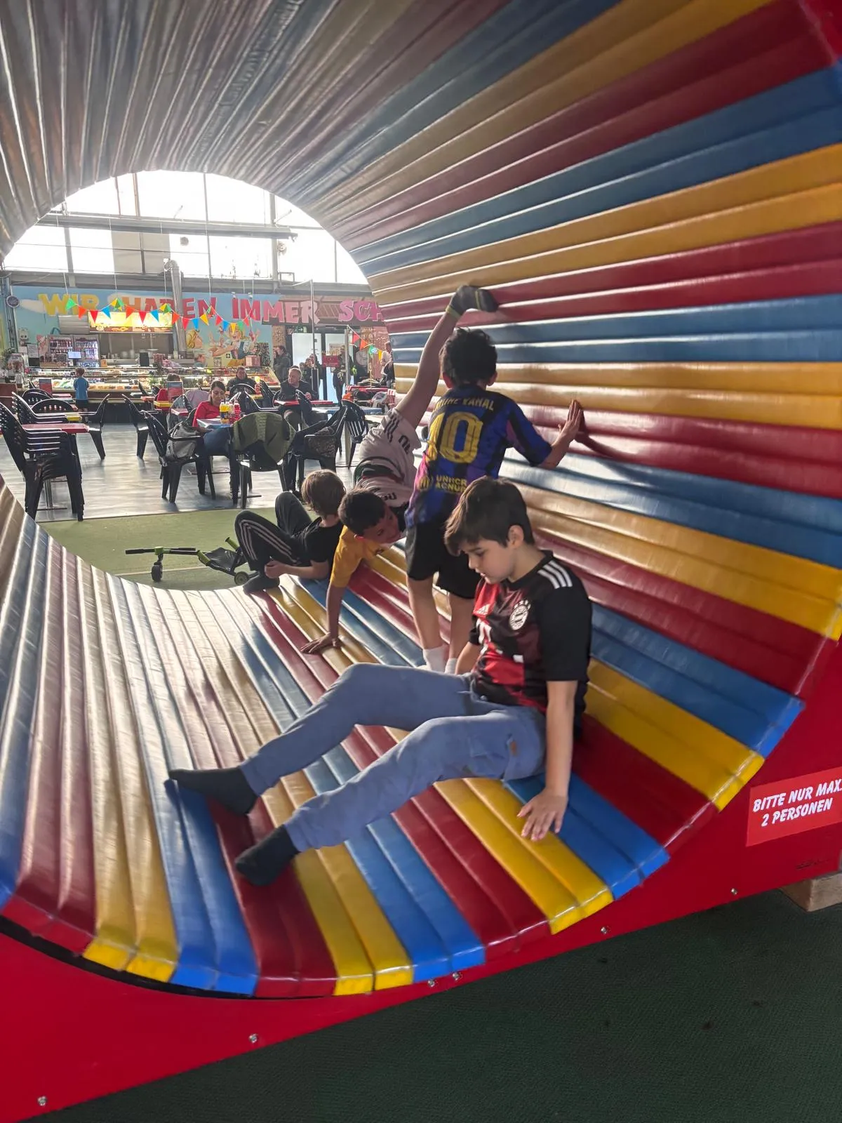 Kinder rutschen und klettern in einem farbigen, aufblasbaren Tunnel, während andere Besucher im Hintergrund sitzen.