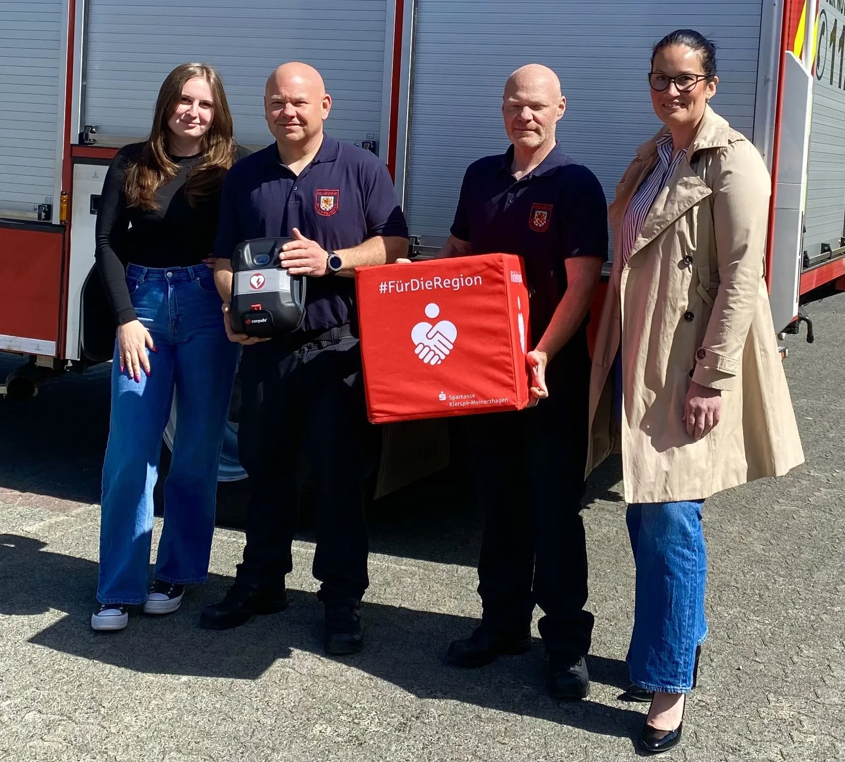 Zwei Frauen der Sparkasse überreichen einen Defibrillator und eine Tasche mit dem Aufdruck #FürDieRegion an zwei Feuerwehrleute vor einem roten Feuerwehrauto.