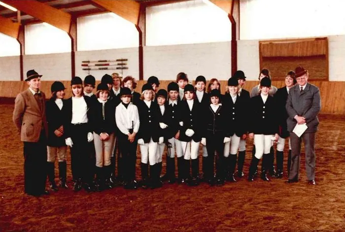 Eine Gruppe von etwa 20 jungen Reitern und Reitern, die in einem Reitstall posieren, flankiert von zwei erwachsenen Männern in formeller Kleidung. Die meisten Reiter tragen schwarze Reitjacken und Helme.