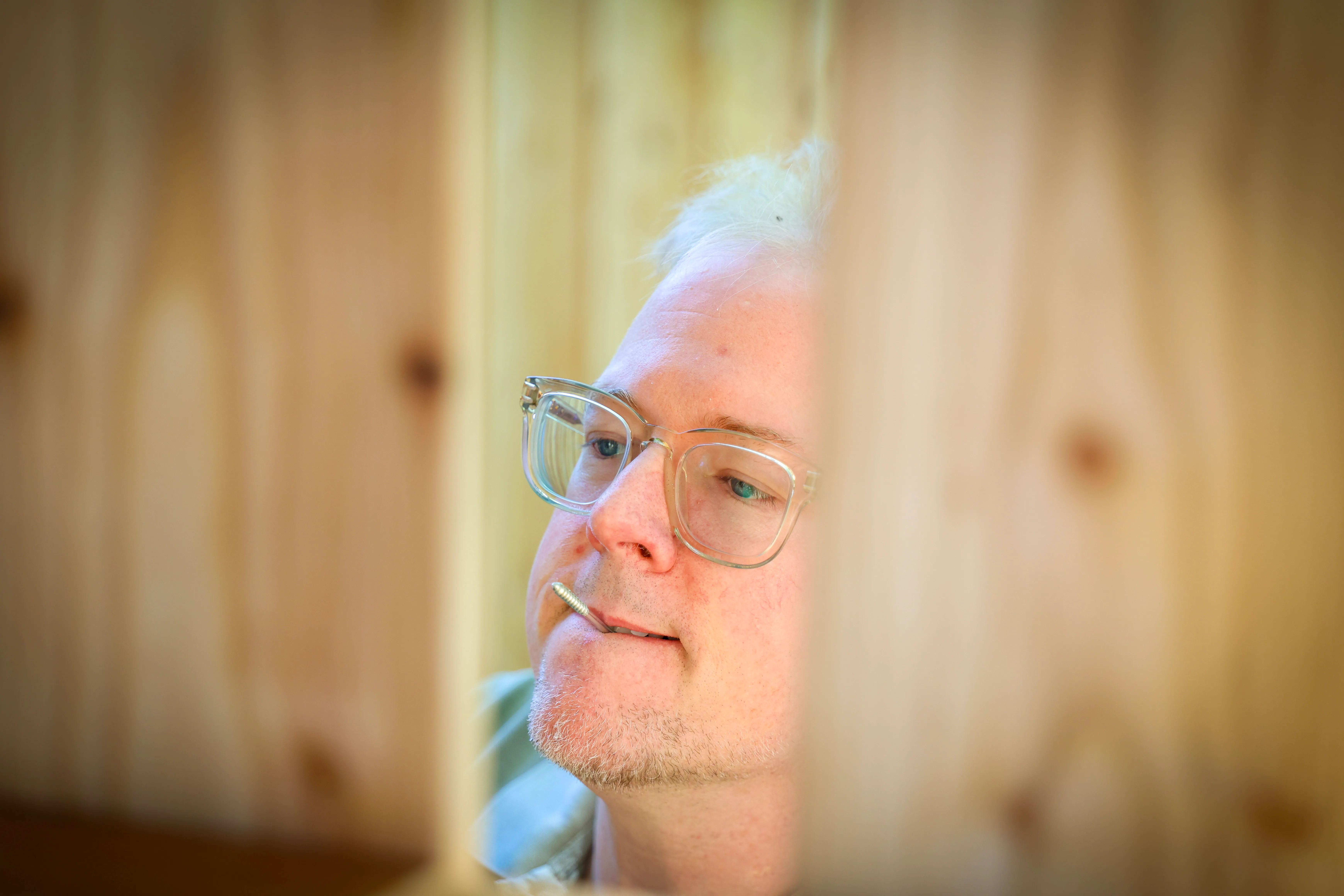 Nahaufnahme eines Mannes mit Brille und weißem Haar, der vor einem Holzpaneel sitzt und in die Ferne blickt.