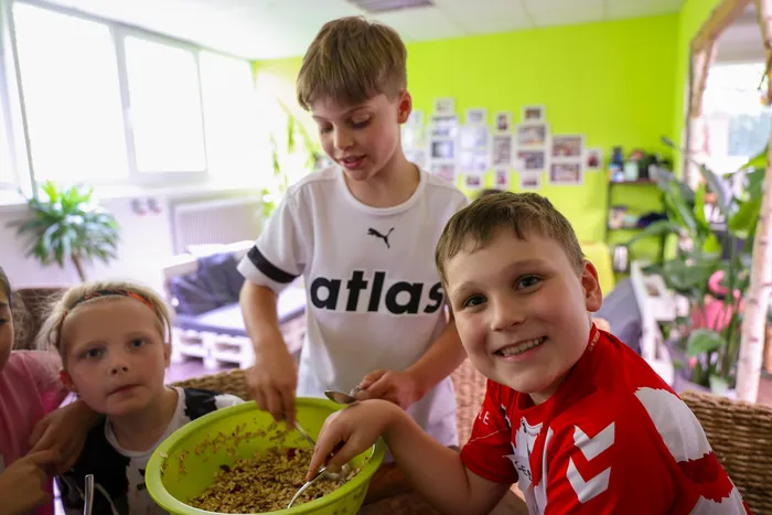 Kinder bereiten in einem hellen Raum gemeinsam eine Mahlzeit zu. Ein Junge in rotem Trikot rührt in einer Schüssel mit braunen Lebensmitteln, während ein anderer Junge im Hintergrund hilft.