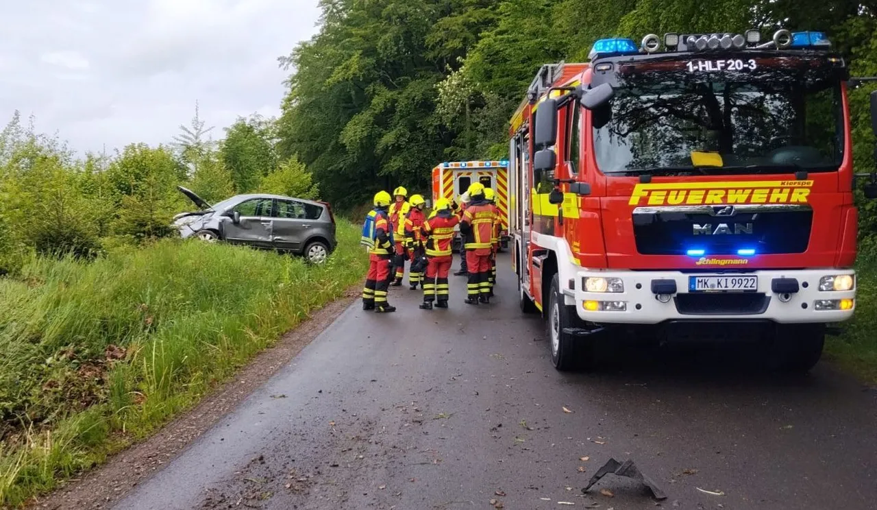 Rotes Feuerwehrauto an einer Landstraße, neben einem beschädigten silbernen PKW. Mehrere Feuerwehrleute in voller Montur stehen am Straßenrand.