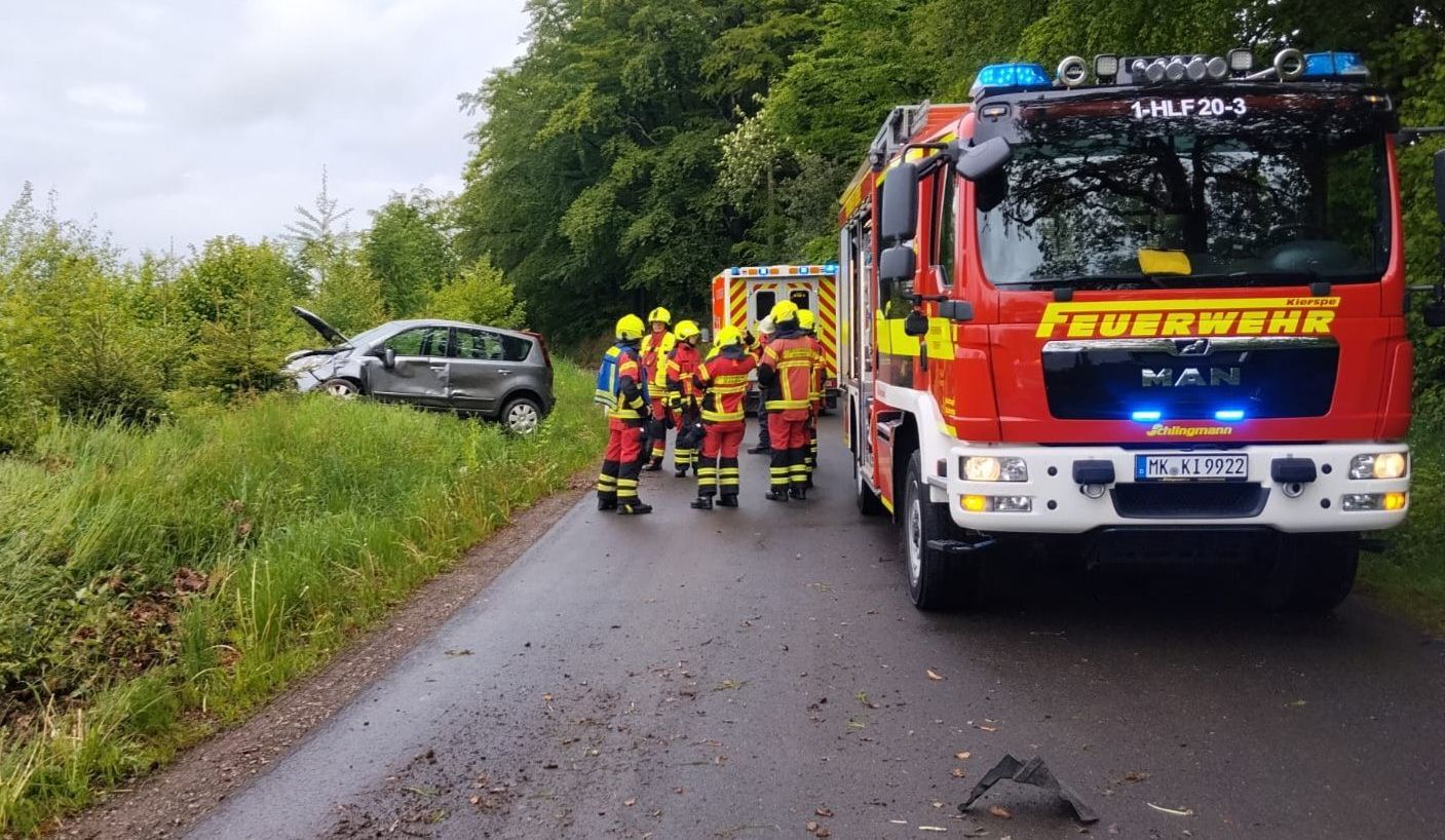Rotes Feuerwehrauto an einer Landstraße, neben einem beschädigten silbernen PKW. Mehrere Feuerwehrleute in voller Montur stehen am Straßenrand.