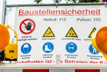 Schild mit Warnhinweisen zur Baustellensicherheit: Zutritt verboten, Absturzgefahr, schwebende Lasten, Elektrizität, Helmpflicht, Sicherheitsschuhe, Schutzbrille und Schutzkleidung.