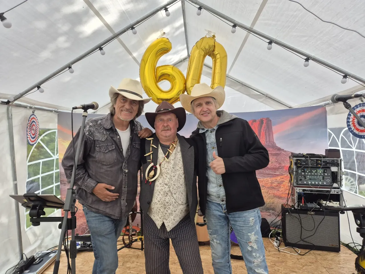 Drei Männer mit Cowboyhüten stehen vor einer Bühne unter einem Zelt. Im Hintergrund eine Kulisse mit Wüstenlandschaft und goldene Ballons mit der Zahl 60.