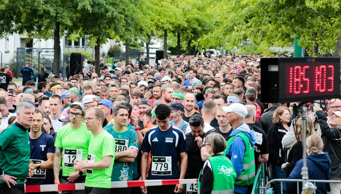 Eine große Menschenmenge läuft an einer roten Barriere entlang, während eine digitale Uhr 18:51:03 anzeigt. Die Läufer tragen Sportkleidung und Startnummern, während Zuschauer am Rand stehen.