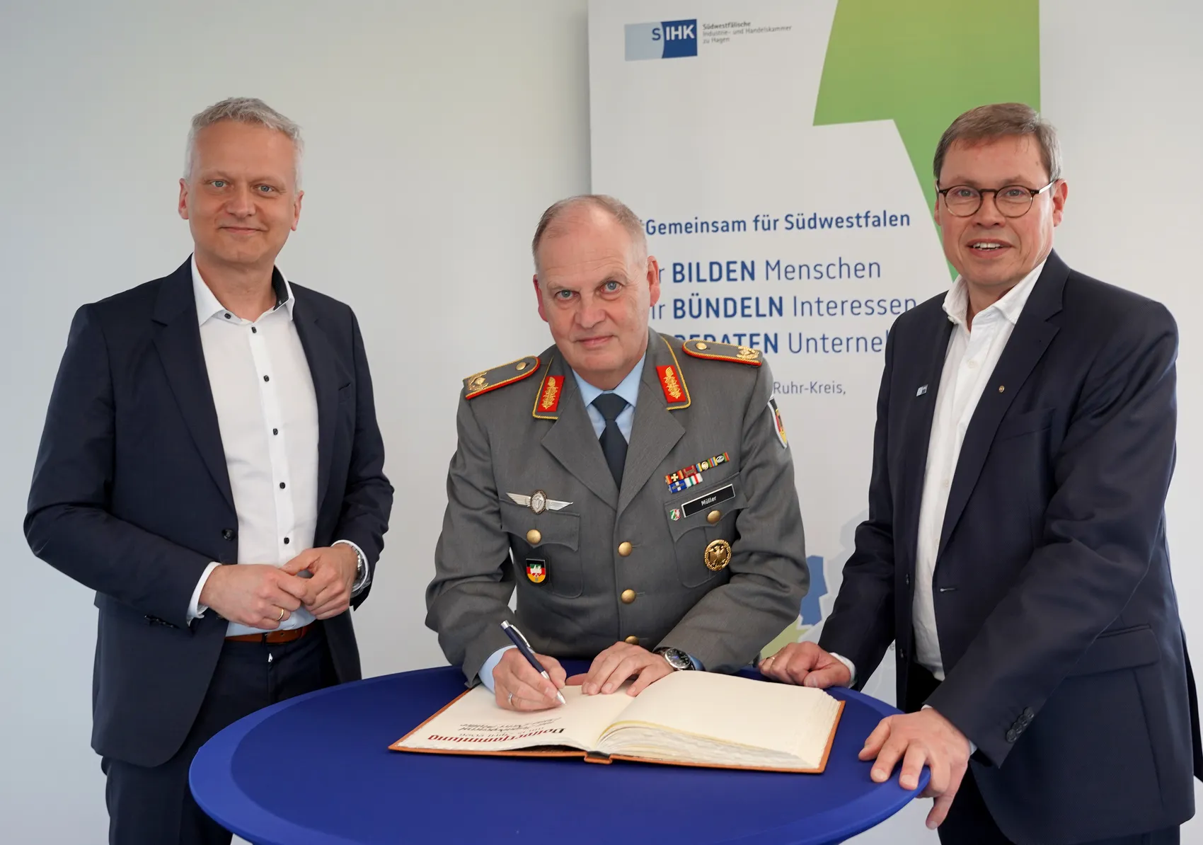 Drei Männer in Anzügen stehen an einem runden Tisch. Der Mann in der Mitte, Brigadegeneral Müller, unterzeichnet ein Dokument.