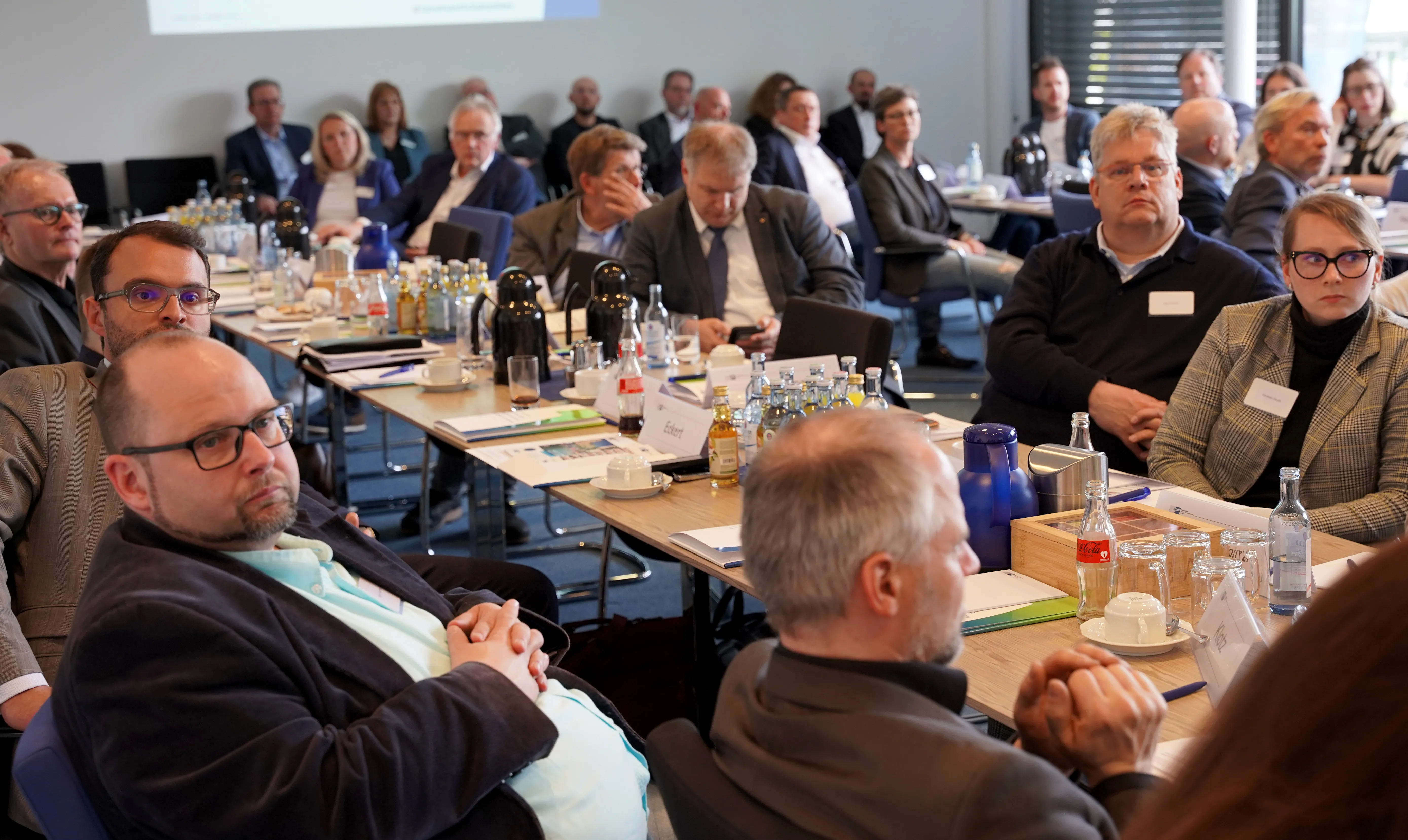 Verschiedene Personen sitzen an Tischen in einem Konferenzraum und verfolgen eine Präsentation im Hintergrund.